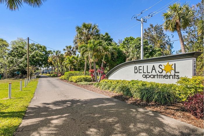 Inviting entrance to Bella Sol Apartments, showcasing lush greenery and a welcoming sign under a clear blue sky.