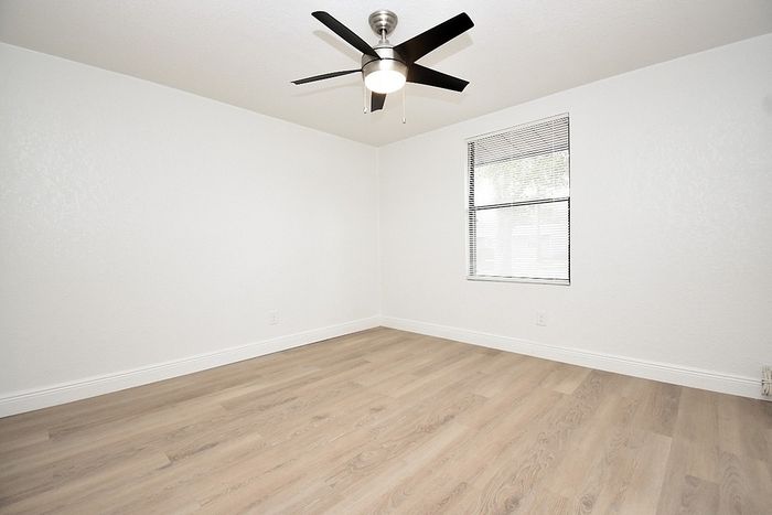 Bright, minimalist bedroom featuring elegant hardwood floors, sleek ceiling fan, and large window for natural light.