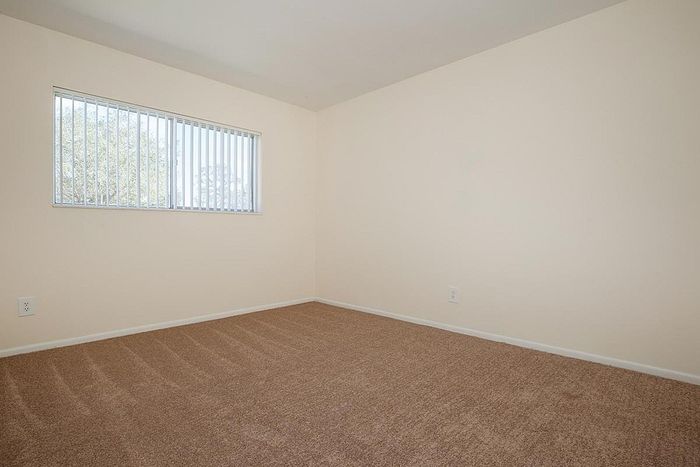 Bright and airy empty room with soft carpet flooring, large window with vertical blinds, and neutral walls, perfect for personalization.