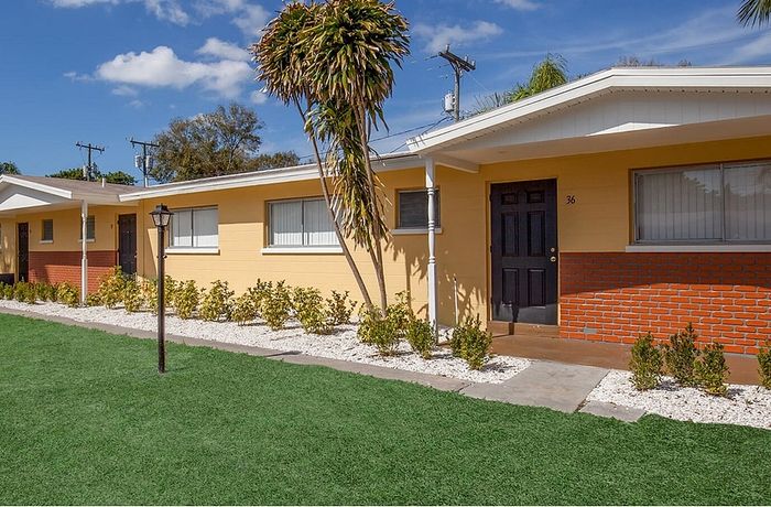 Charming single-story apartment exterior with yellow walls, brick accents, and inviting landscaping under a bright blue sky.