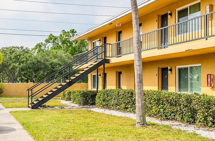 Charming two-story apartment building with yellow exterior, black railings, and lush green landscaping.