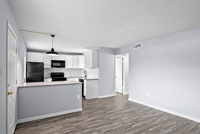 Modern apartment interior featuring a sleek kitchen with white cabinetry, granite countertops, and stainless steel appliances, accompanied by a spacious open living area with elegant wood flooring.