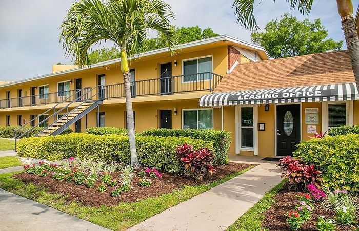 Charming apartment complex with vibrant landscaping and a welcoming leasing office, complete with tropical palm trees and manicured hedges.