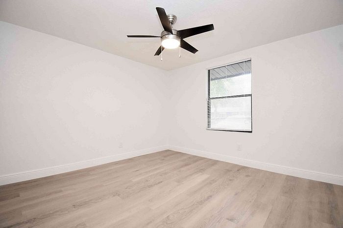 Bright and spacious room with elegant wood flooring, modern ceiling fan, and natural light streaming through a sleek window.