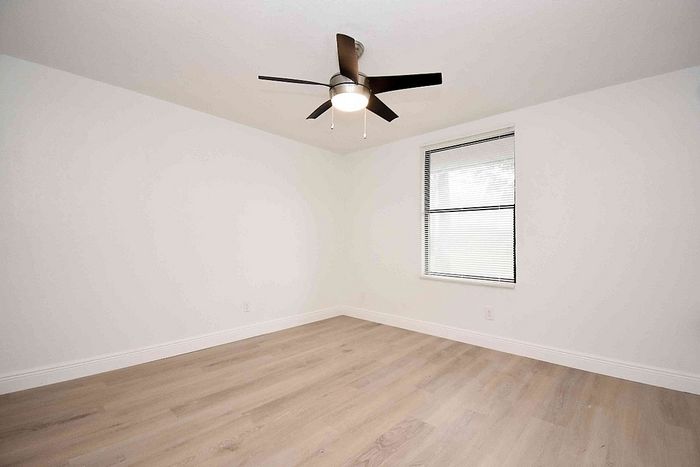 Bright, airy bedroom with sleek ceiling fan and large window, featuring pristine white walls and elegant wood flooring.