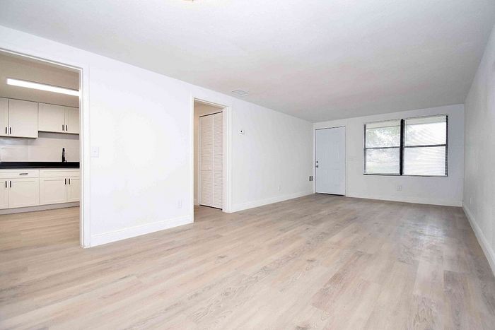 Bright and spacious living area with elegant light wood flooring, large window, and modern kitchen access.