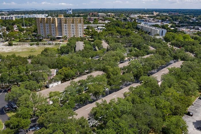 Aerial view of a vibrant urban neighborhood with lush greenery, featuring a tall apartment building amidst tree-lined streets and residential areas.