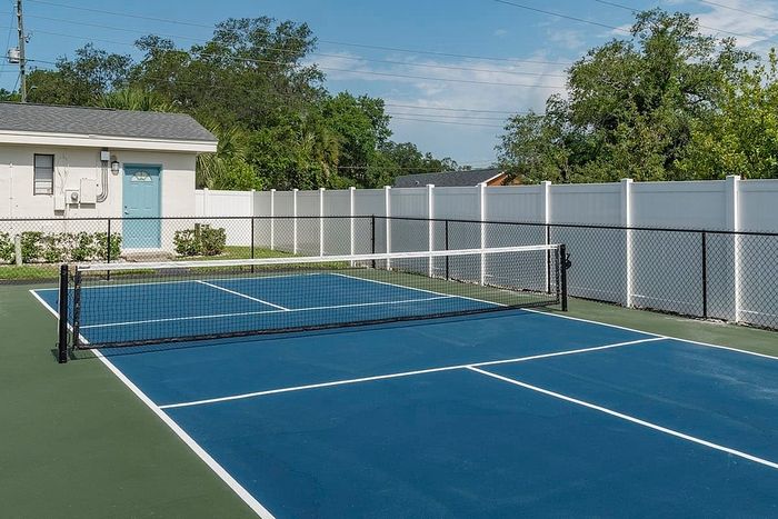 Modern outdoor tennis court with fresh blue and green surfaces, surrounded by a white privacy fence and lush greenery.