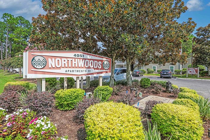 Charming and inviting entrance to Northwoods Apartments II, surrounded by lush greenery and vibrant flowers under a clear blue sky.