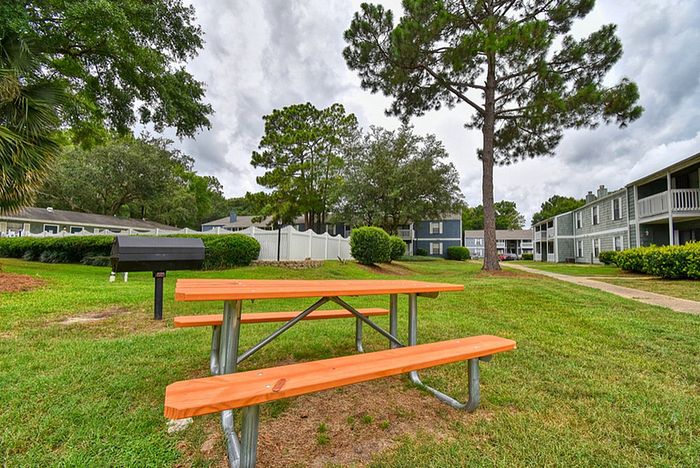 Inviting outdoor space with a wooden picnic table and grill, surrounded by lush greenery and charming apartment buildings.