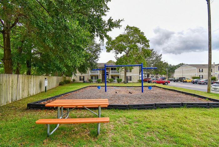 Charming residential community featuring a vibrant playground with swings and a picnic table, surrounded by lush greenery and cozy apartment buildings.