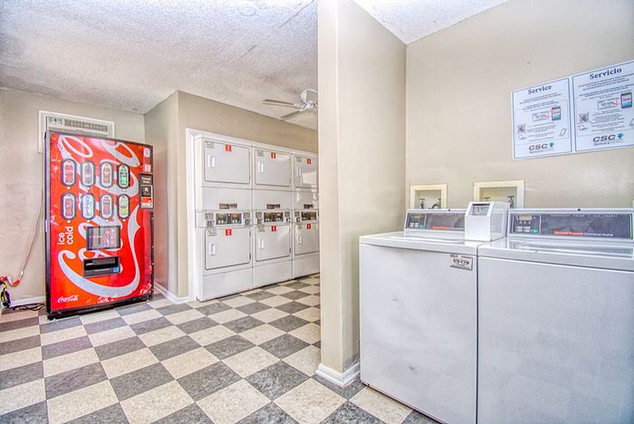 Brightly lit laundry room featuring modern washers and dryers adjacent to a convenient soda vending machine, set on a stylish checkered tile floor.