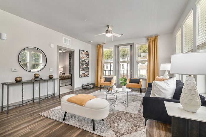 Studio Apartments in Birmingham, AL - Tributary Rise - Living Room With Hardwood-Style Flooring, Area Rug, Couch, End Tables With Lamps, Armchairs, Bench, Coffee Table, Ceiling Fan With Light, Display Shelf, Round Mirror, Windows With Blinds, And Glass Door With Access To Patio.