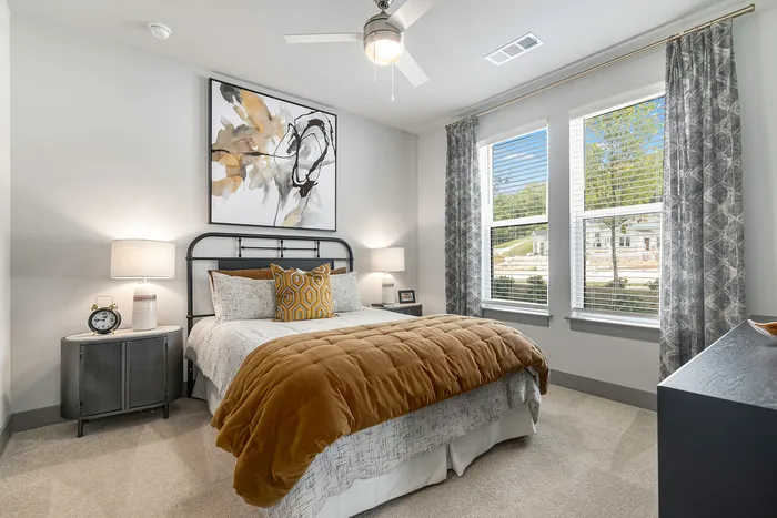 Two-Bedroom Apartments in Birmingham, AL - Tributary Rise - Bedroom with Carpet Flooring, Bed, Nightstands with Lamps, Wall Art, Windows with Blinds and Curtains, Ceiling Fan, and Dresser.