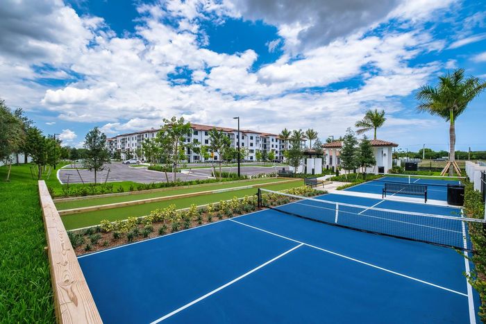 Cooper City Senior Living - Two Tennis Courts Outside Of Apartment Homes. Courts Have Bench And Trash Can On Site. Surrounded By Palms And Landscape.