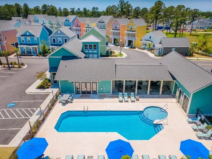 Myrtle Beach, SC, Pet-Friendly Luxury Apartments - Isle Cottages - Aerial View of Resort-Style Pool With Sundeck, Blue Umbrellas, Lounge Chairs, Clubhouse Patio, and Colorful Cottage-Style Homes.
