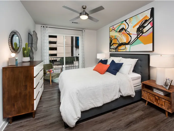 1-Bedroom Apartments in Midtown Atlanta, GA - Spectrum on Spring - Furnished Bedroom with Wood-Style Plank Flooring, Bed, Wooden Nightstand with Lamp, Wooden dresser with White Details, Mirror, Ceiling Fan, Armchair, and Sliding Glass Door with Access to Balcony