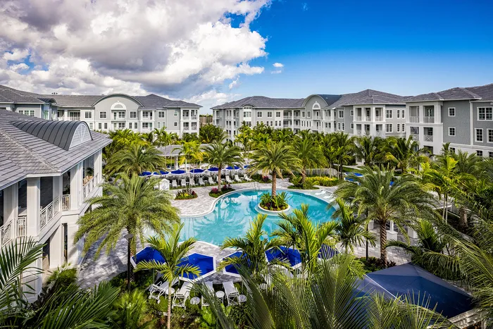 Pet-Friendly Apartments in Lantana - Manor Lantana - Resort-Style Pool with Umbrella Lounge Chairs, Palm Trees, and Apartment Complex View