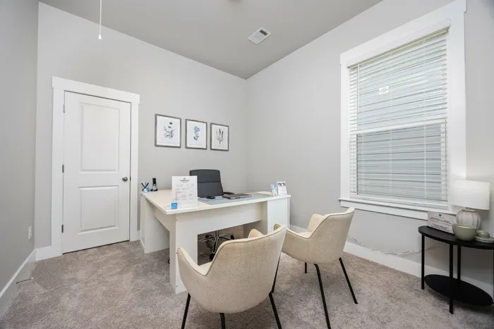 Luxury Homes in Opelika, AL - Champions Square - Home Office with a Desk, Office Chair, Two Tan Seating Chairs, Plush Carpeting, and a Large Window with White Blinds.