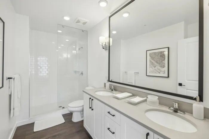 Studio Apartments in Lantana, FL - Ivory Lantana - Bathroom with Wood-Style Flooring, Dual Sinks, Quartz Countertop, Large Vanity Mirror, and Glass-Walled Walk-In Shower.