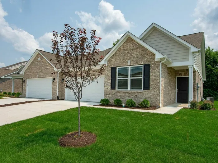 Two-Bedroom Homes for Rent in Avon, IN - Woods at Countryside Crossing - Home Exterior with Beige Bricks, Black Door and Window Shutter Details, Two-Car Garage, Driveway, Landscaped Plant Beds, Front Lawn, and Tree.
