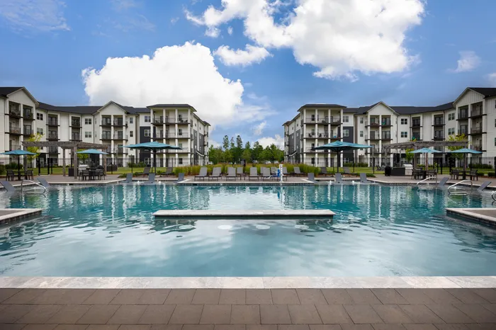 1-Bedroom Apartments in Athens, AL - Gateway Village at Swan Creek - Resort-Style Swimming Pool With Sundeck Lounge Chairs, Umbrellas, and Modern Apartment Buildings Under Blue Sky.