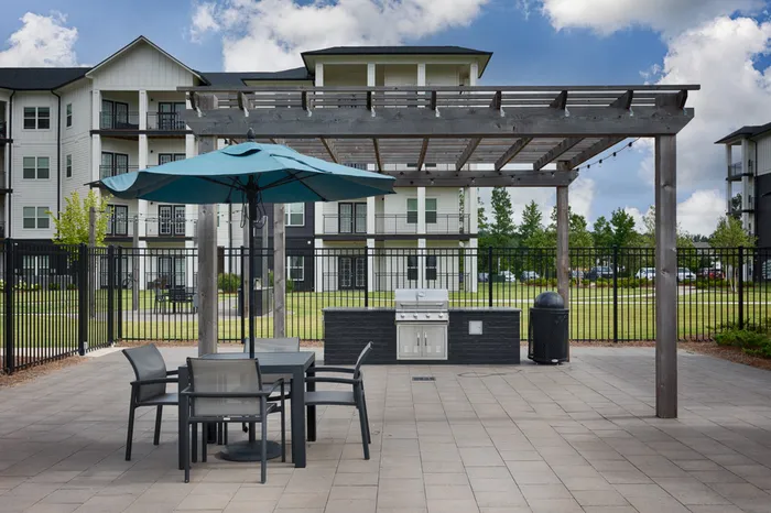 One-Bedroom Apartments in Athens, AL - Gateway Village at Swan Creek - Outdoor Grilling Patio With Pergola, Gas Grill Station, Bistro Seating, Blue Umbrella, and Fenced Courtyard Views.