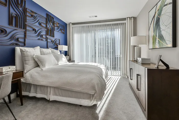 One-Bedroom Luxury Apartments in Sandy Springs, GA - The Morgan Sandy Springs - Bedroom with Carpet Flooring, Queen Bed, Desk, Chair, Dresser, Lamps, Wall Decor, and a Sliding Glass Door.