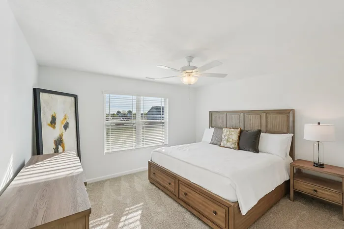 Two-BR Homes in Whitestown, IN - Prairie Chase - Bedroom With Carpet Flooring, Large Bed with Built-In Storage, Nightstand, Lamp, Ceiling Fan With Lighting, Oversized Painting, And Dresser.