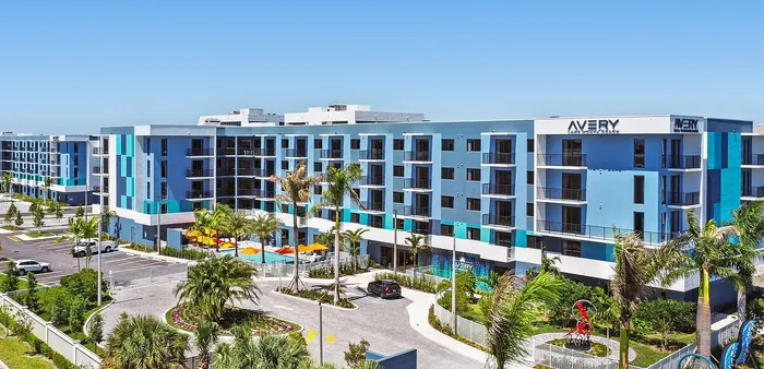 Modern blue apartment complex with spacious balconies, surrounded by lush greenery and palm trees, featuring a vibrant central courtyard with orange umbrellas and a lively pool area.