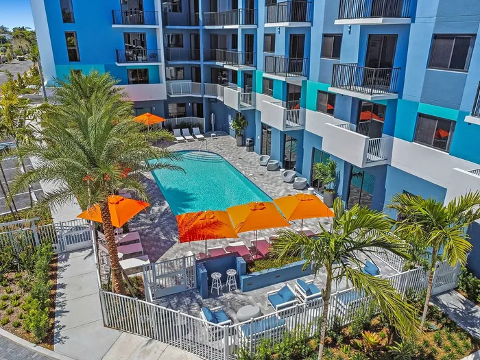 Vibrant apartment complex with a stunning outdoor pool, surrounded by palm trees and modern seating, offering an inviting oasis for relaxation.