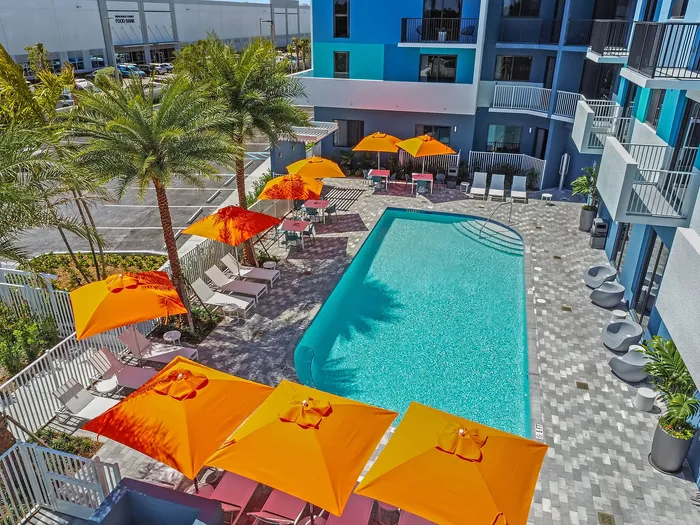 Vibrant apartment courtyard with a refreshing pool, surrounded by lush palm trees and bright orange umbrellas, creating a tropical oasis vibe.