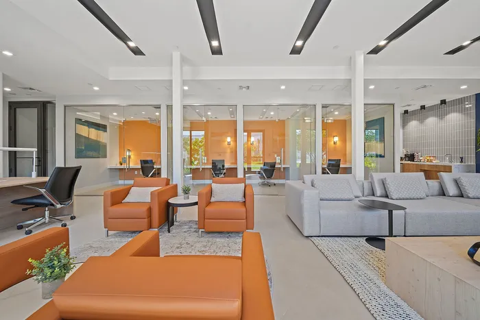 Modern meeting area with stylish orange and gray furnishings, glass walls, and a kitchenette, offering a chic and inviting atmosphere.