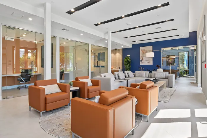 Modern and inviting lounge area featuring cozy orange chairs, a sleek glass-enclosed office space, and stylish decor with blue accent walls.