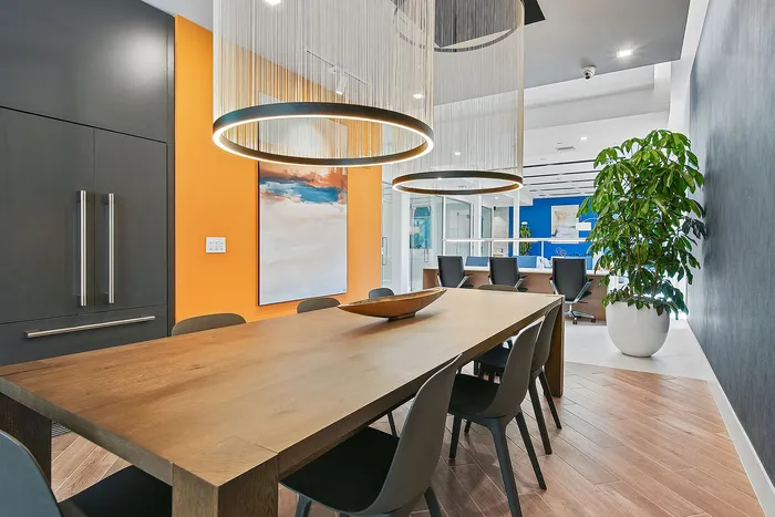 Modern dining area with a sleek wooden table, contemporary lighting fixtures, vibrant orange accent wall, and lush greenery enhancing the elegant atmosphere.