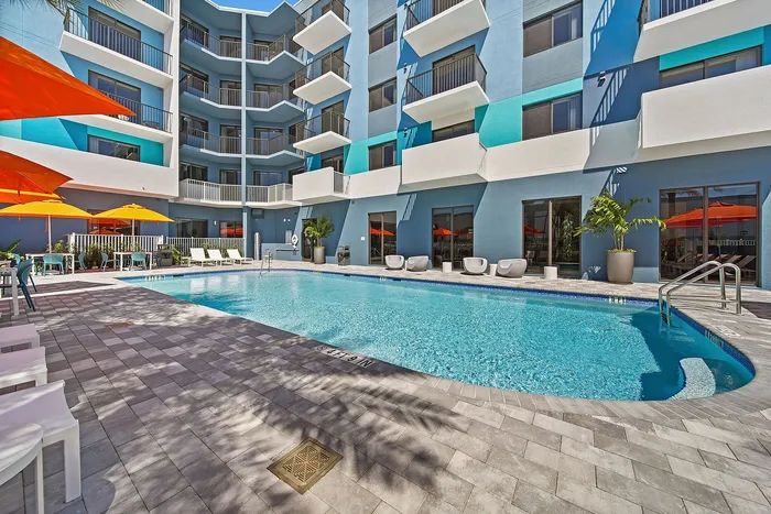 Luxury apartment community featuring a pristine outdoor pool, surrounded by modern blue facade and vibrant orange umbrellas, perfect for relaxation and leisure.