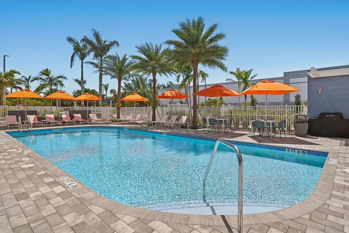 Inviting apartment pool area with sparkling blue water, surrounded by palm trees and orange sun umbrellas providing a tropical retreat.