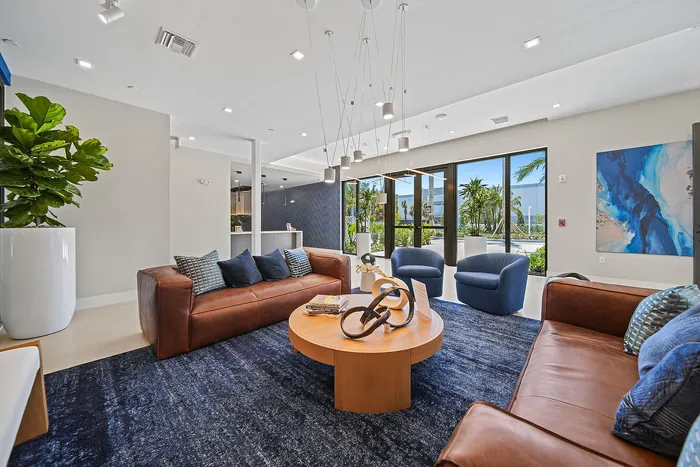 Modern clubroom area with chic brown leather sofas, elegant blue armchairs, and a stylish wooden coffee table, overlooking lush greenery through expansive windows.
