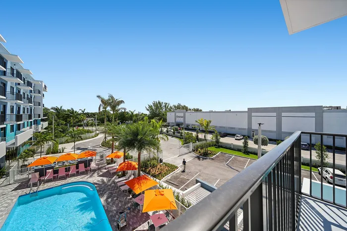 Vibrant apartment complex with a sparkling swimming pool, surrounded by lush palm trees, offering a relaxing outdoor space with bright orange umbrellas and comfortable lounge chairs