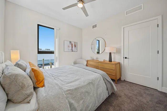 Inviting bedroom with a modern design, featuring a cozy bed, elegant dresser, and ample natural light through a large window.