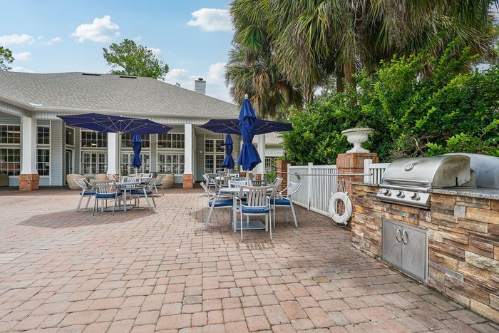 Inviting outdoor patio with dining tables under blue umbrellas, a built-in BBQ grill, and lush greenery, perfect for entertaining and relaxation.