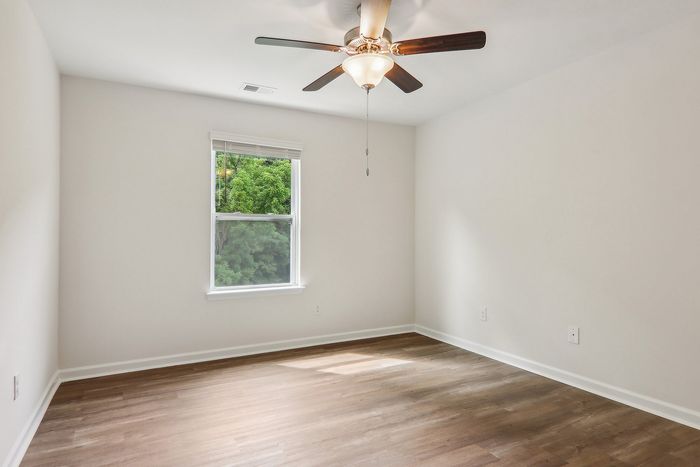 Bright and airy room with elegant wood flooring, large window with lush outdoor view, and modern ceiling fan.