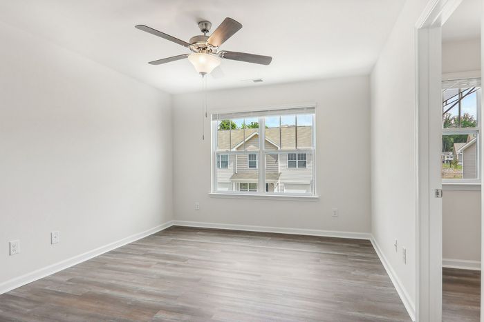 Bright and airy bedroom with elegant hardwood floors, large window, and ceiling fan, offering a tranquil suburban view.