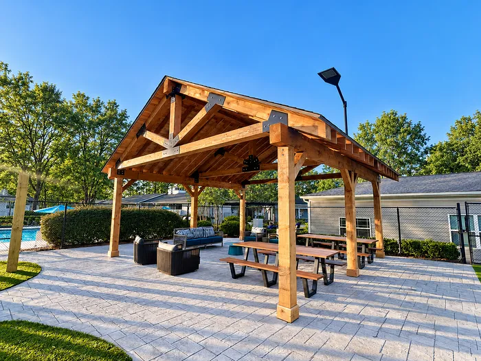 Charming outdoor pavilion with wooden beams and comfortable seating, perfect for gatherings; adjacent to a sparkling pool surrounded by lush greenery.