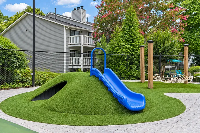 Charming outdoor playground with a vibrant blue slide on a grassy hill, surrounded by lush greenery and modern apartments.