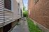 Charming urban walkway nestled between a well-maintained home and rustic brick building, leading to a lush green garden.