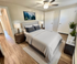 Elegant bedroom with modern decor, plush bedding, stylish ceiling fan, and warm hardwood floors, enhanced by natural lighting.