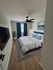 Elegant bedroom with cozy bedding, modern art, and natural light streaming through the window. Features sleek wooden flooring, a large ceiling fan, and stylish blue accents.