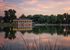 Serene lakeside pavilion with glowing lights reflecting on tranquil water at sunset, surrounded by lush greenery.