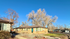 Charming single-story brick home with large windows, surrounded by mature trees and set against a bright blue sky.